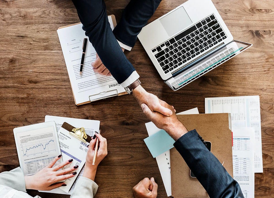 Two people shake hands across a desk full of documents and a laptop