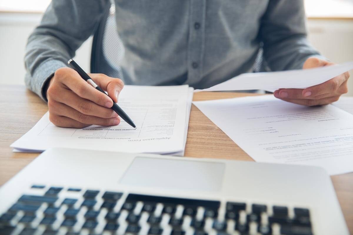 Close-up of documents on a desk, with a person sitting in front of them comparing a tabular list against a document held in their hand