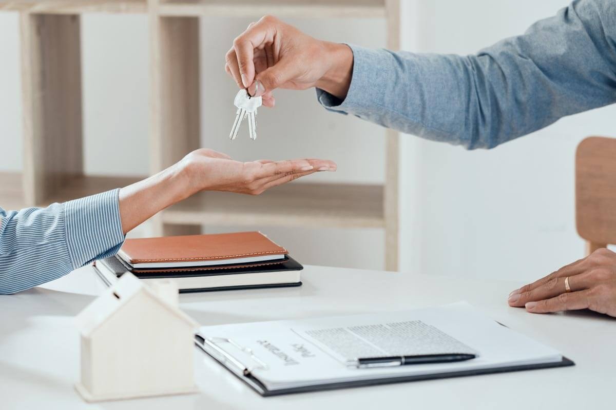 Close-up of a key handover over a desk with a clipboard holding documents, two notebooks, and a house-shaped piggy bank