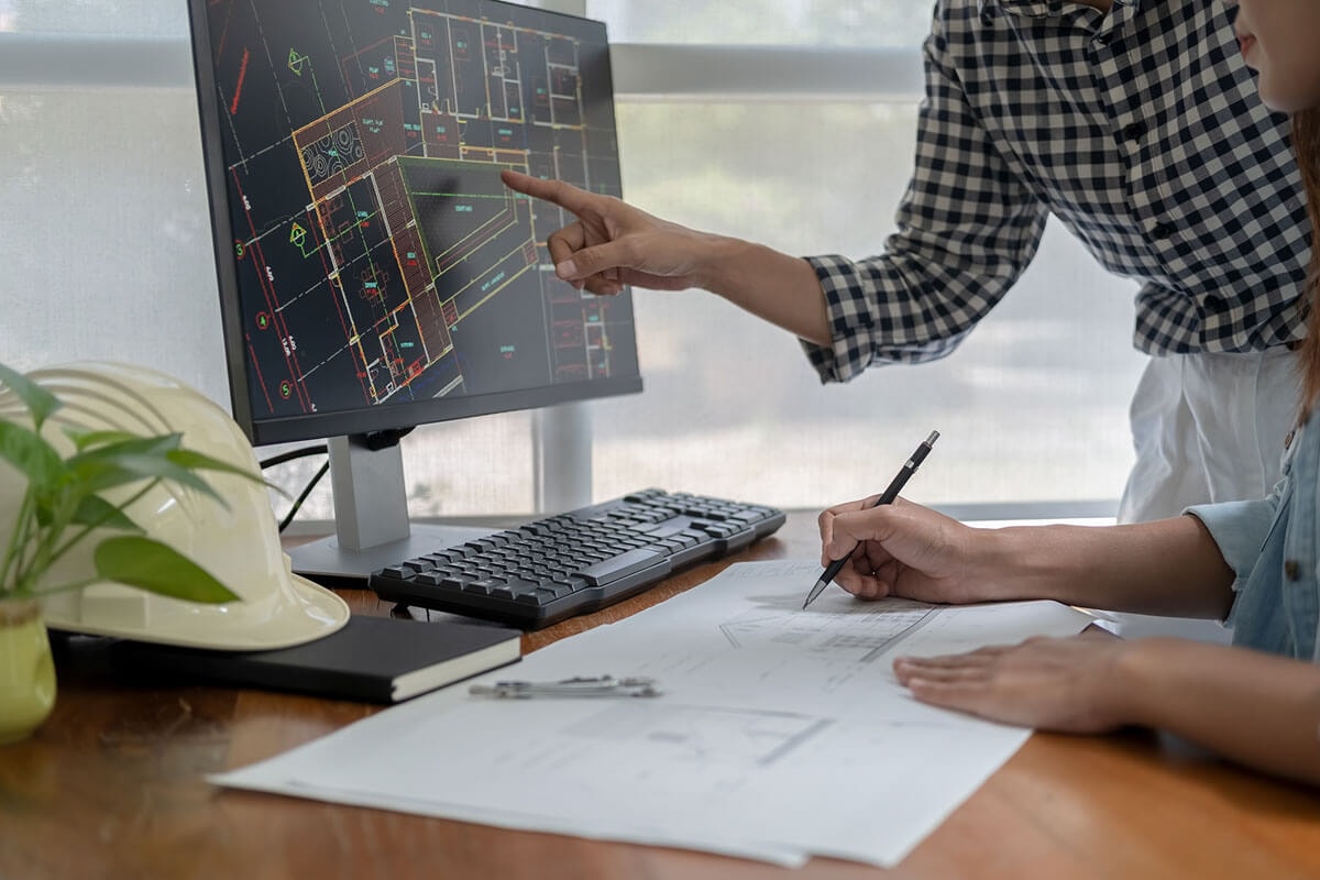 Two people at a desk with a PC, one pointing to a detail in the blueprint on the monitor while the other transfers something with a pen onto the facade elevation drawing in front of them.