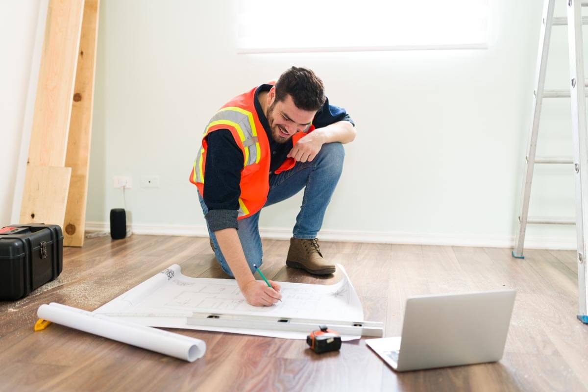 Interior fitting specialist kneels on a wooden floor in front of a floor plan and takes notes on it