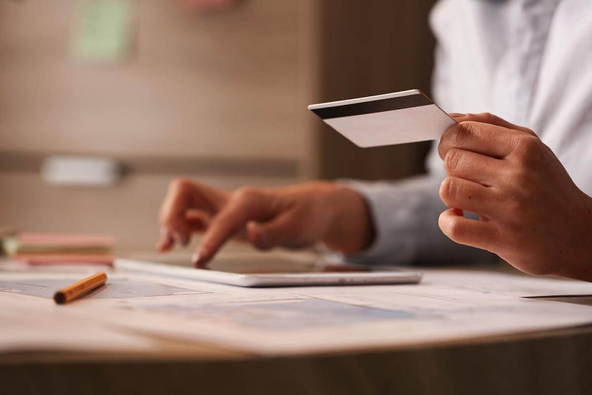 Left hand holding a credit card while the right hand types on a tablet in the background