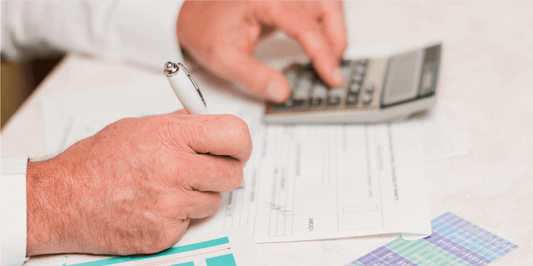 Close-up of two hands — one entering information into a form with a ballpoint pen, the other typing numbers into a calculator
