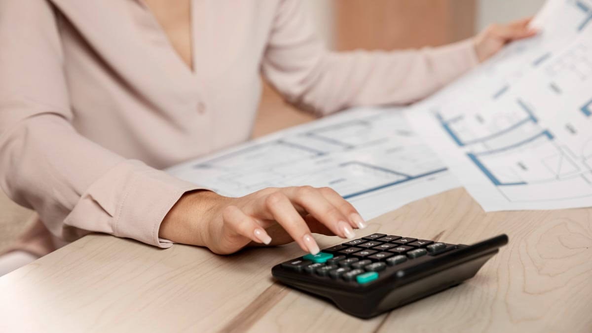 Close-up of a woman's hand at a desk typing on a calculator, with another hand in the background holding up a floor plan halfway