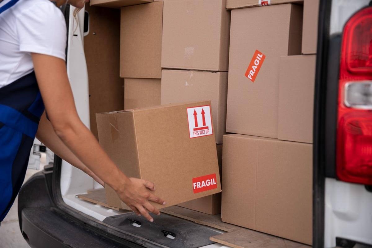 Close-up: a person in blue workwear removes a box labelled with a Fragile sticker from a van; in the background, more stacked boxes fill the loading area.