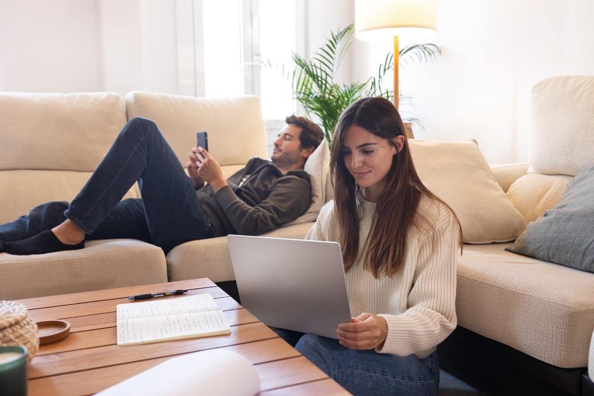 A woman sits on the floor between the sofa and a coffee table working on a laptop, a notebook and pen lie in front of her, and in the background a man lies relaxed on the sofa looking at his smartphone.