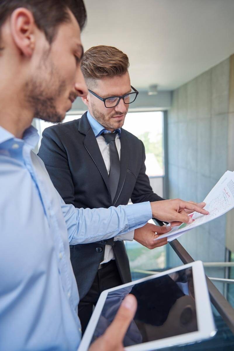 Two men standing on a gallery, going through the details of documents together that one of them is holding.