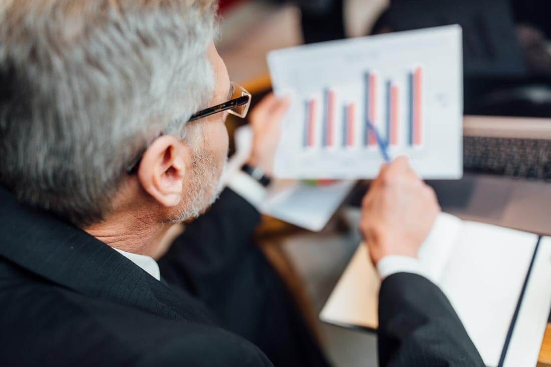 Over-the-shoulder view of an older investment banker sitting at a conference table, reviewing a bar chart.