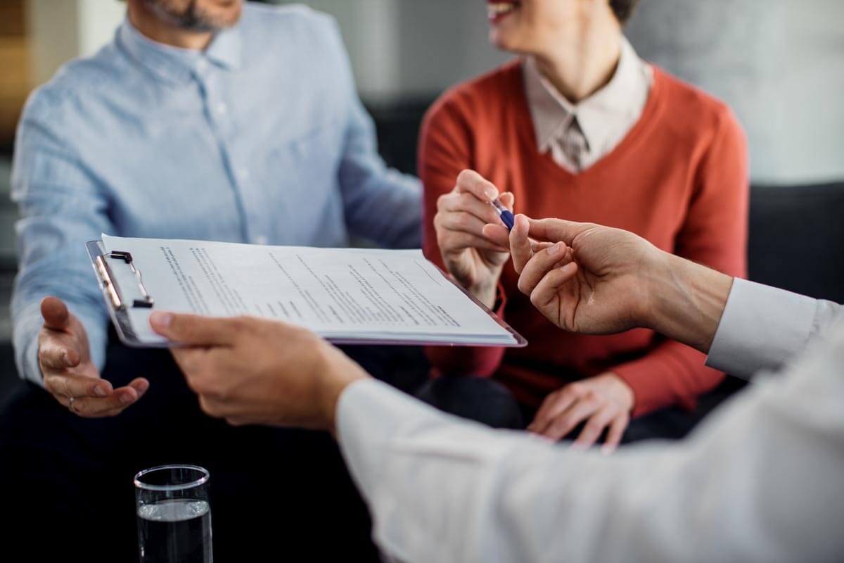 Close-up of a person handing contract documents on a clipboard and a pen to a smiling couple.