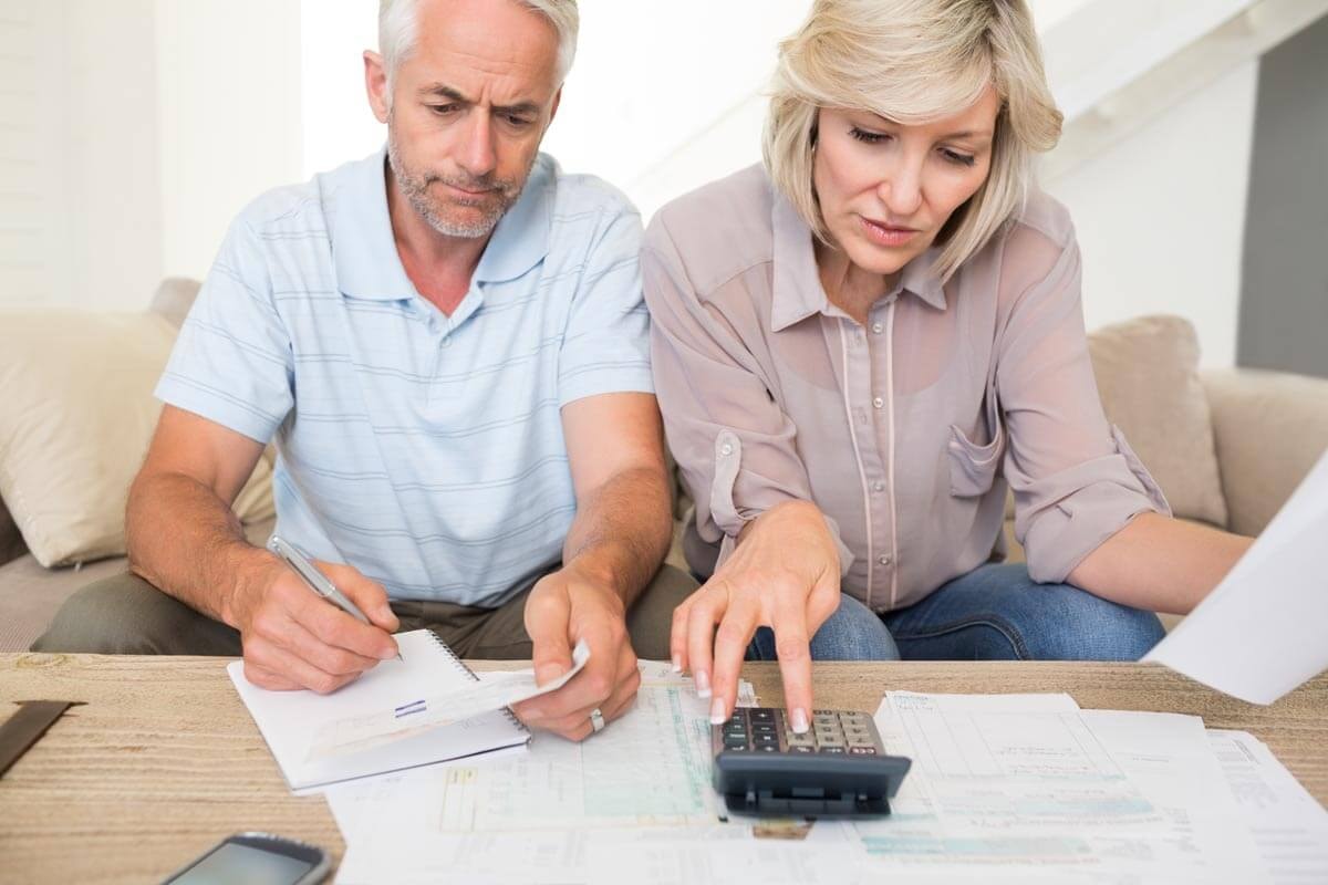A couple sits side by side on a couch in front of a coffee table covered with documents, working through some calculations — she with a calculator, he by hand on a notepad