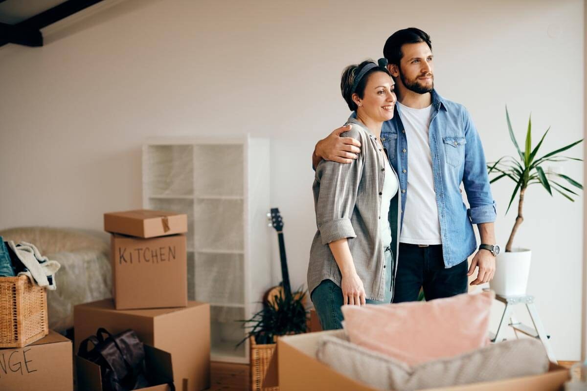 A couple stands contentedly smiling among several open moving boxes and half-assembled furniture.