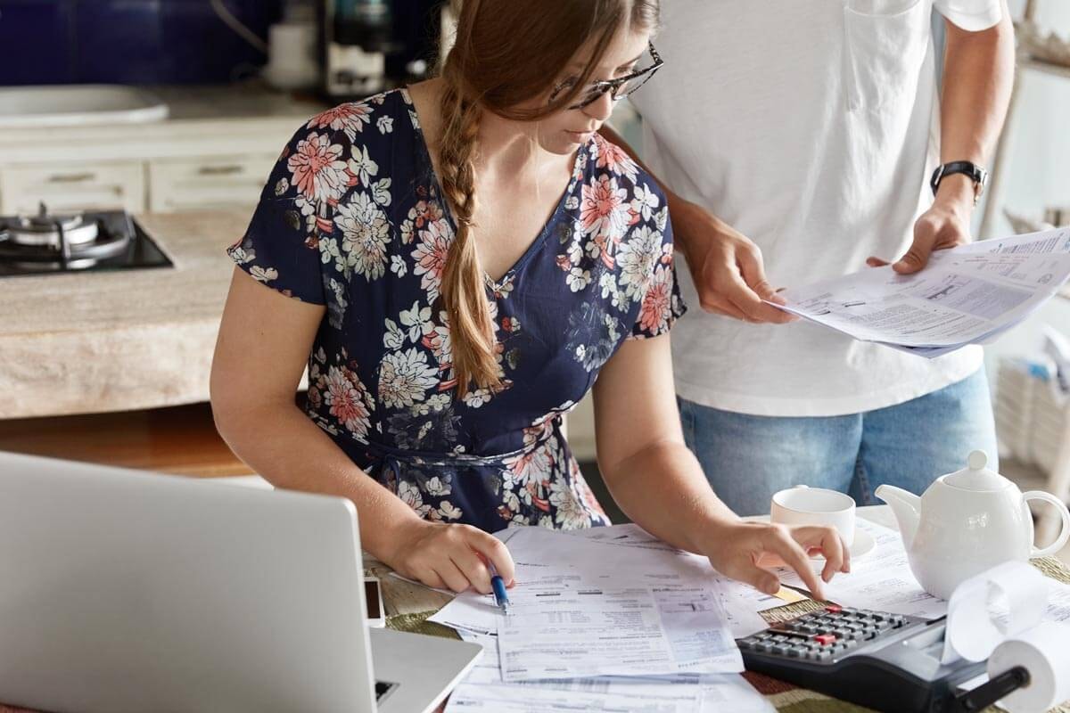A woman sits at a desk making calculations with a calculator, taking information from a stack of documents in front of her on the table, while a man stands next to her holding more documents ready