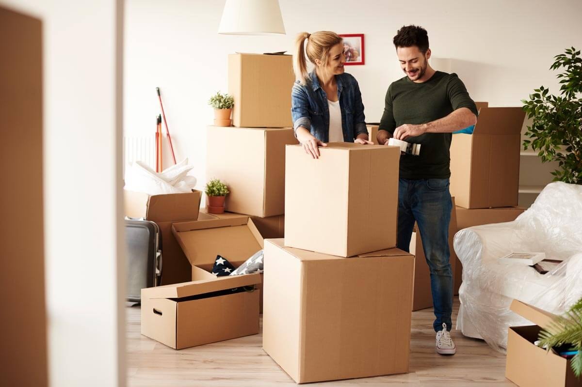 A young couple smiling as they pack moving boxes together; the man is taping up a box while the woman leans against it smiling — surrounded by many boxes, a plastic-wrapped armchair, and houseplants.