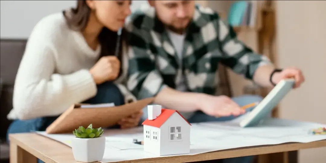 Close-up of a small house miniature and a succulent holding down the edge of a building plan on a table, while in the background a couple sitting on the couch in front of the plan ponder over a document