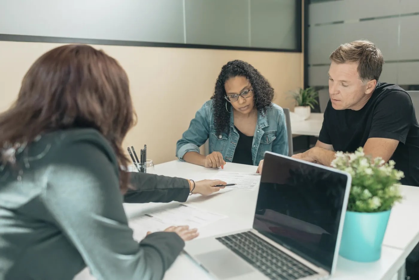 A customer advisor sits at a desk with a laptop, talking to a couple while pointing at the documents between them.