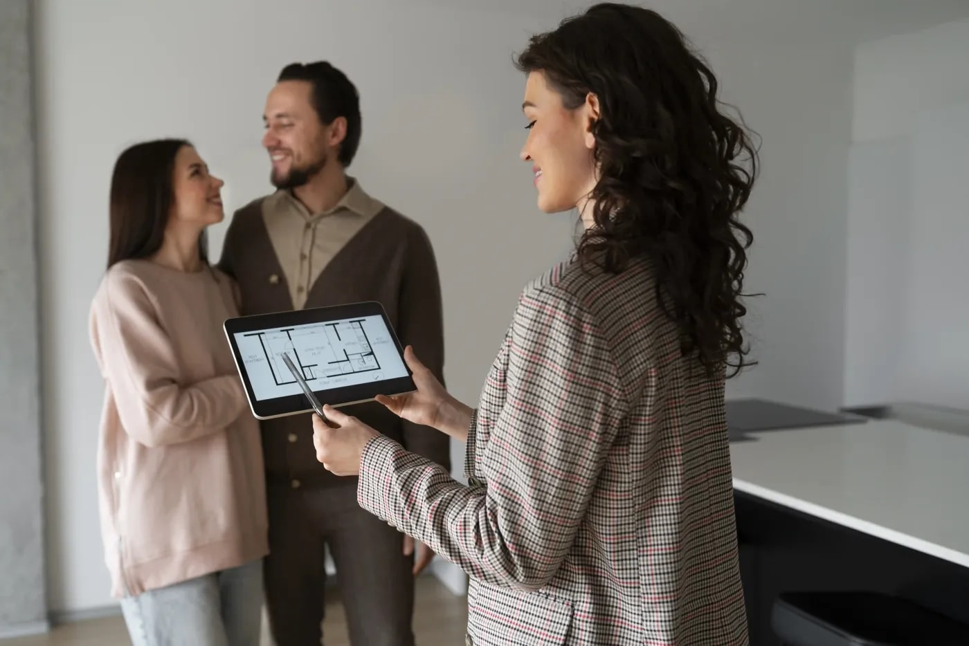 A female real estate agent smiles at a tablet displaying a floor plan, while a couple arm in arm beams happily in the background.