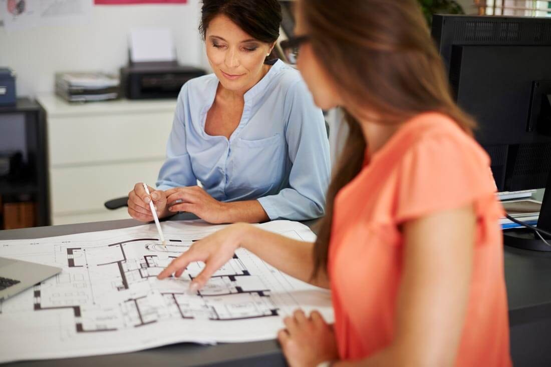Two women sitting opposite each other at a desk, discussing a building plan lying between them on the table, each pointing to a detail.