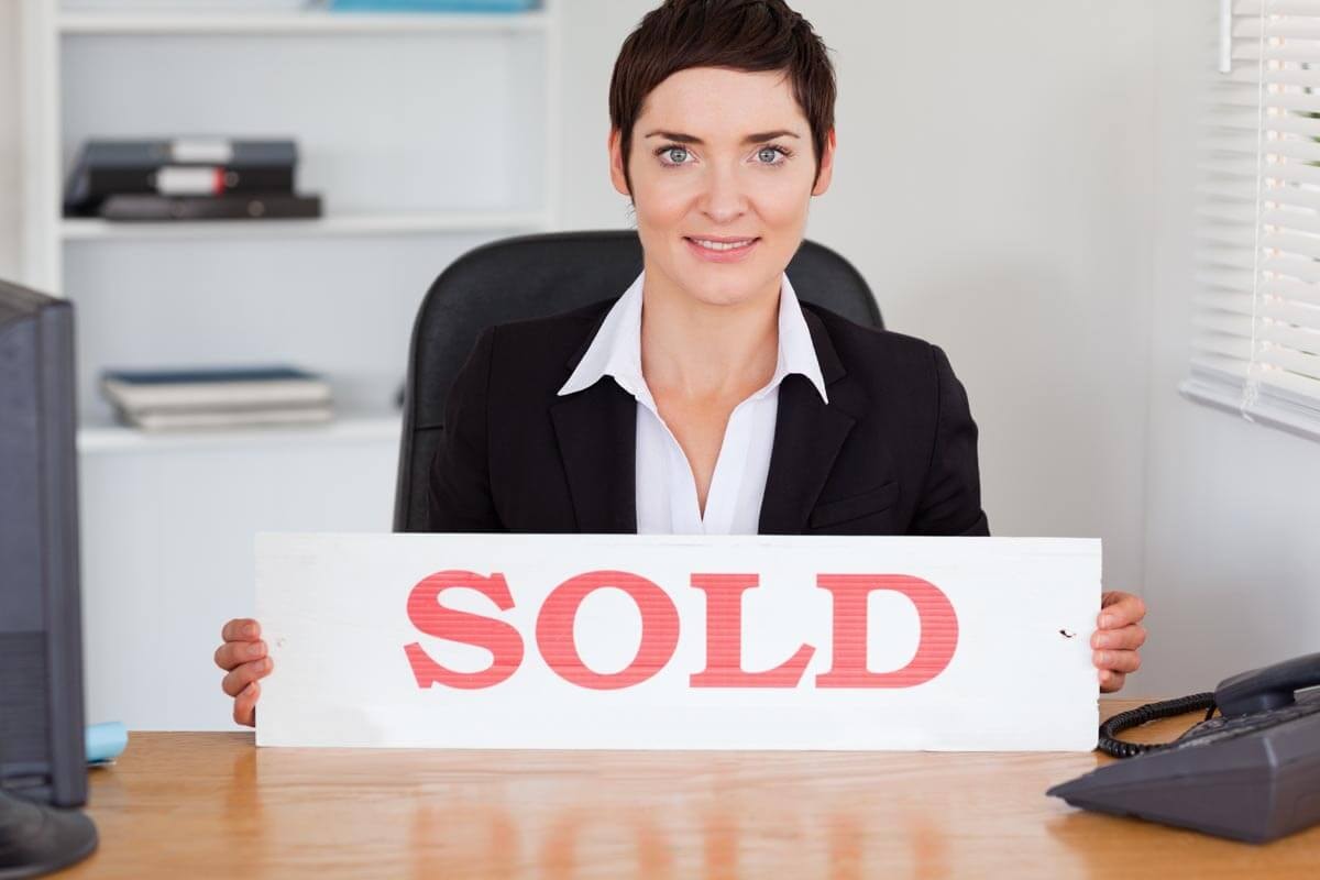 A real estate agent sits at a wooden desk with a PC and phone, holding up a white sign with red lettering that reads 'Sold'