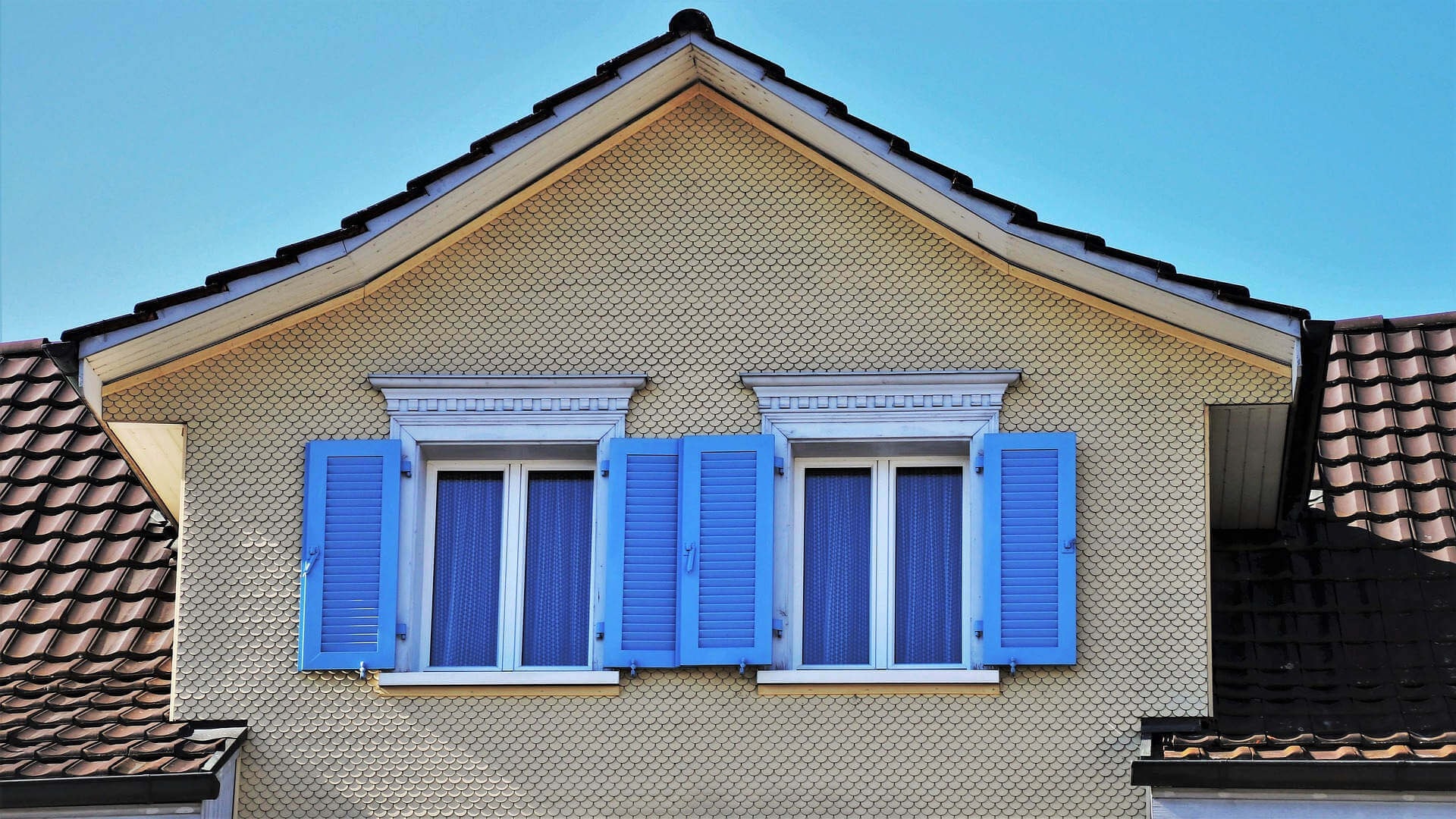 Nahaufnahme einer Dachgaube mit zwei Fenstern und blauen Fensterläden, beigem Wandputz und rotbrauen Dachschindeln vor einem blauen Himmel.