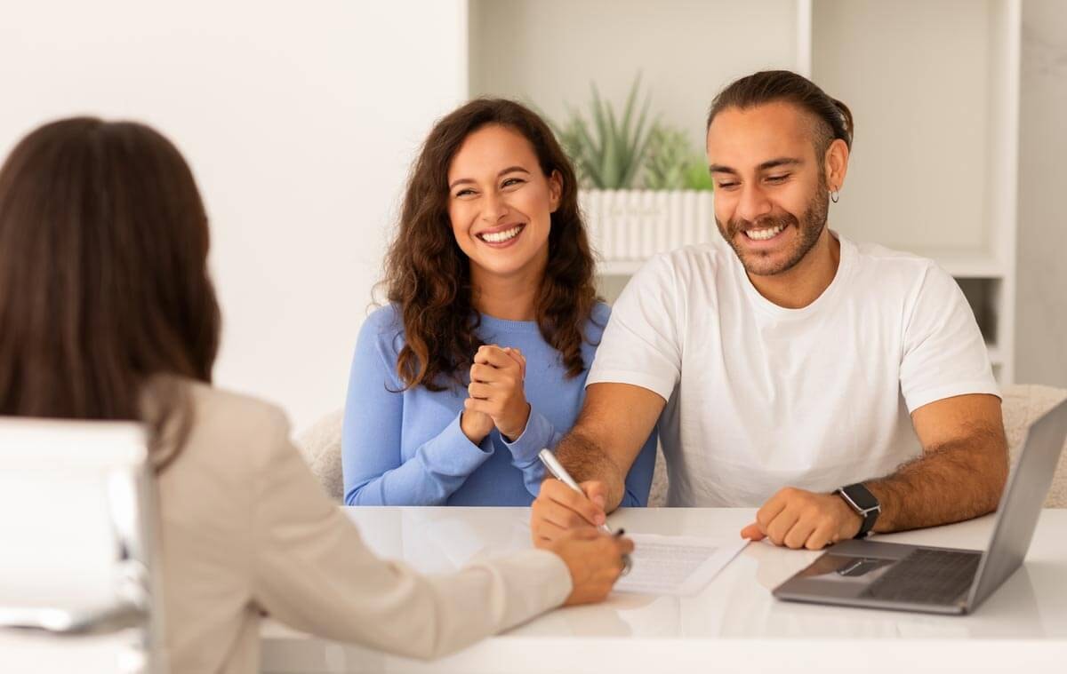 A radiant young couple sits at a table with a female estate agent and joyfully signs contract documents together.