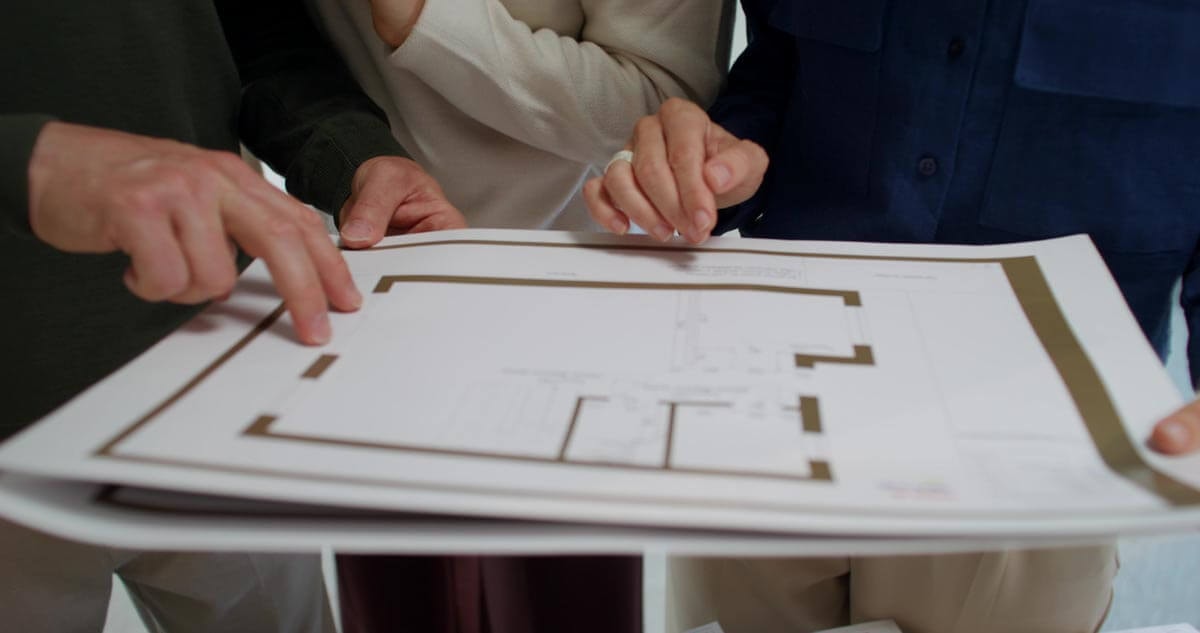 Three people stand around a floor plan, all three holding it up together in the air