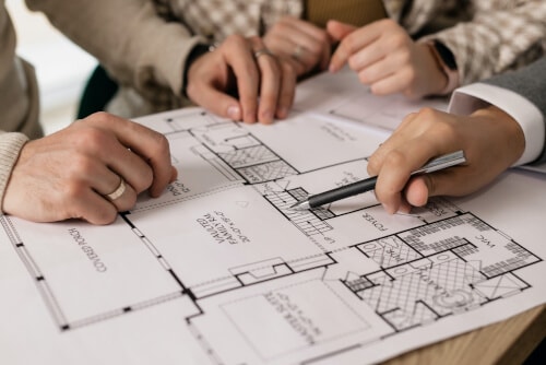 Close-up of a real estate agent with clients over a floor plan, the agent holding a ballpoint pen and pointing to the staircase with it.