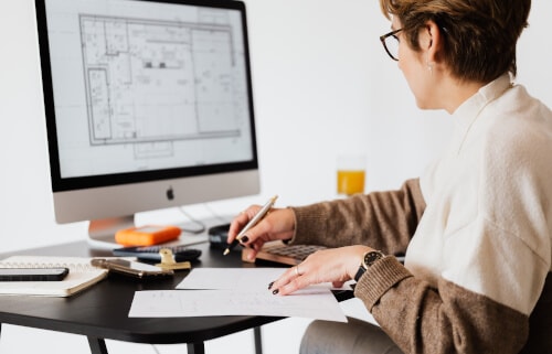 An older woman sits at a desk making handwritten notes about a floor plan shown on her screen