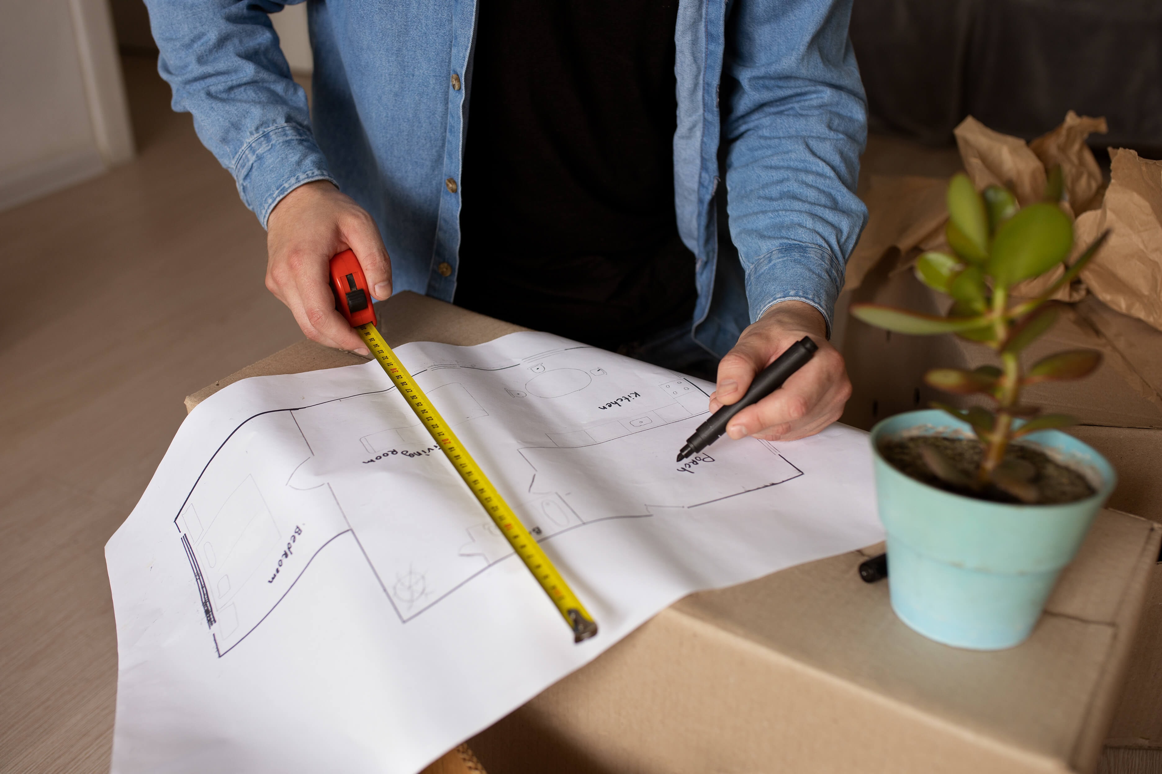 Close-up of a person using a measuring tape and a black pen to add to a rough floor plan lying on a moving box, with a green plant beside it.
