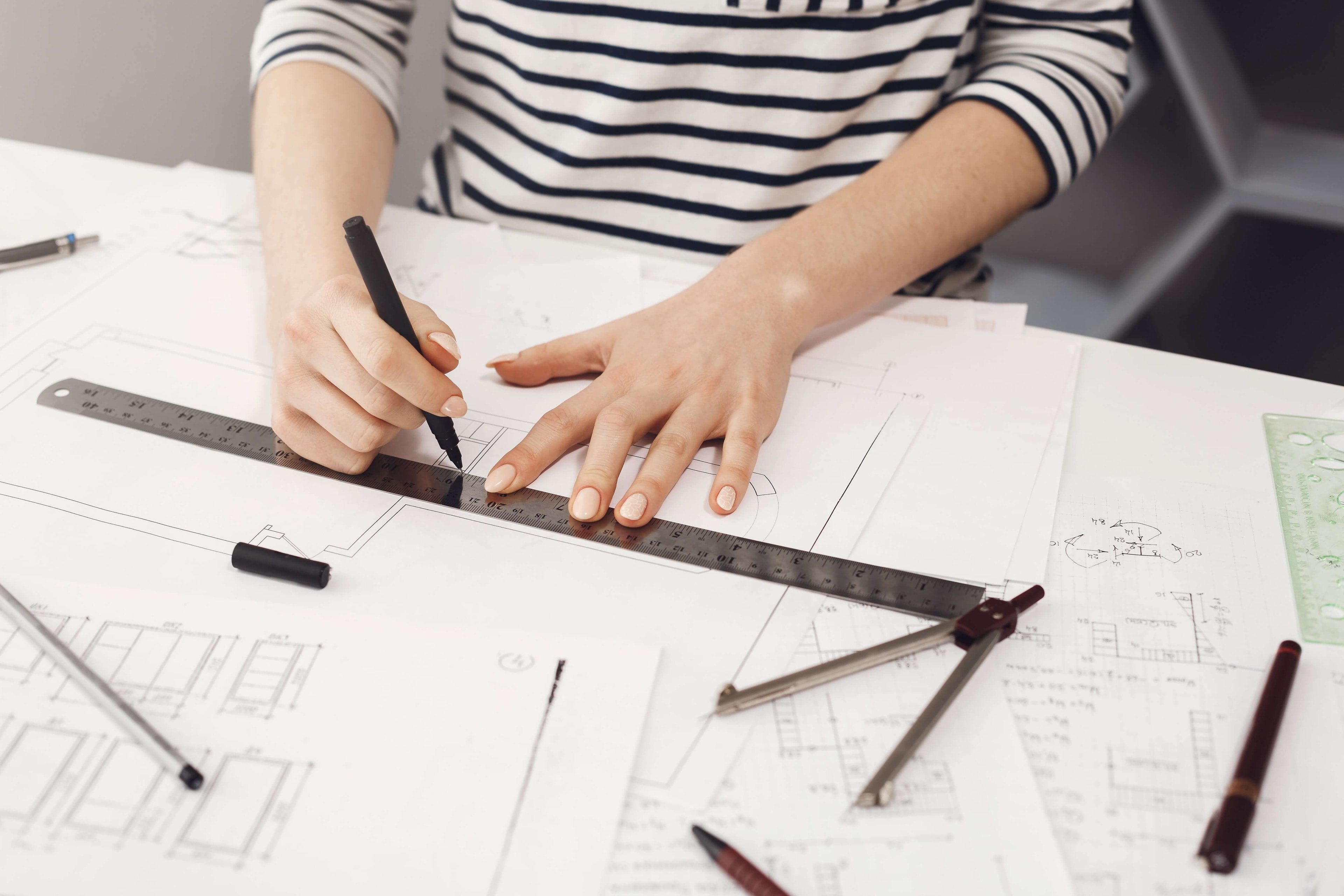 A person draws a floor plan on large paper with a black pen and metal ruler, with several drawing utensils scattered on it.