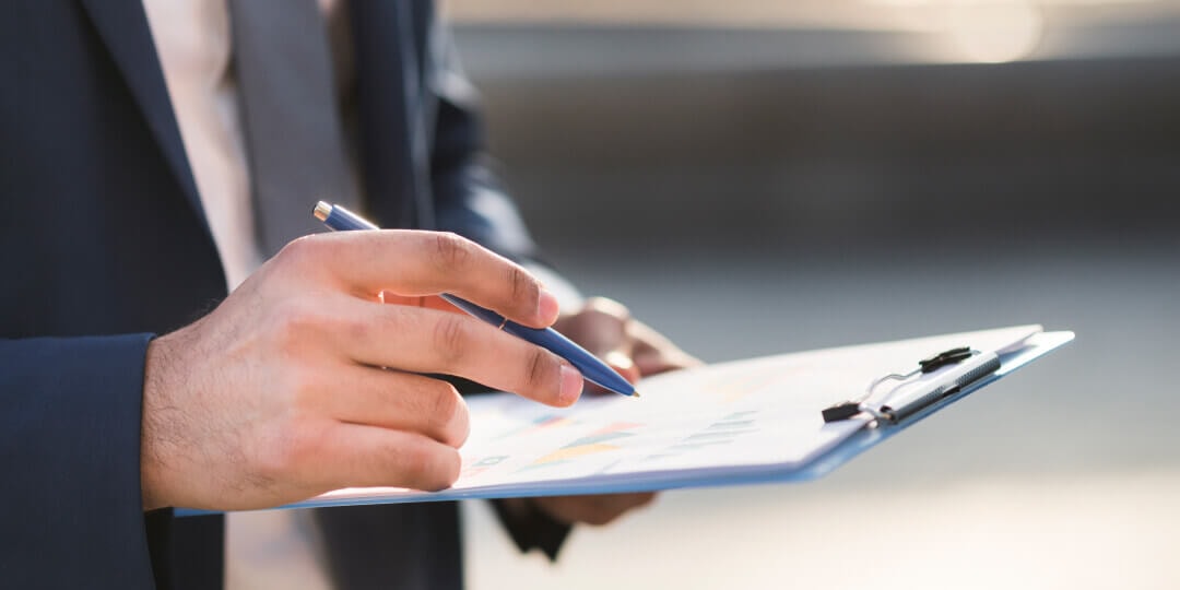 Hand holding a pen over a clipboard with documents and colourful charts.