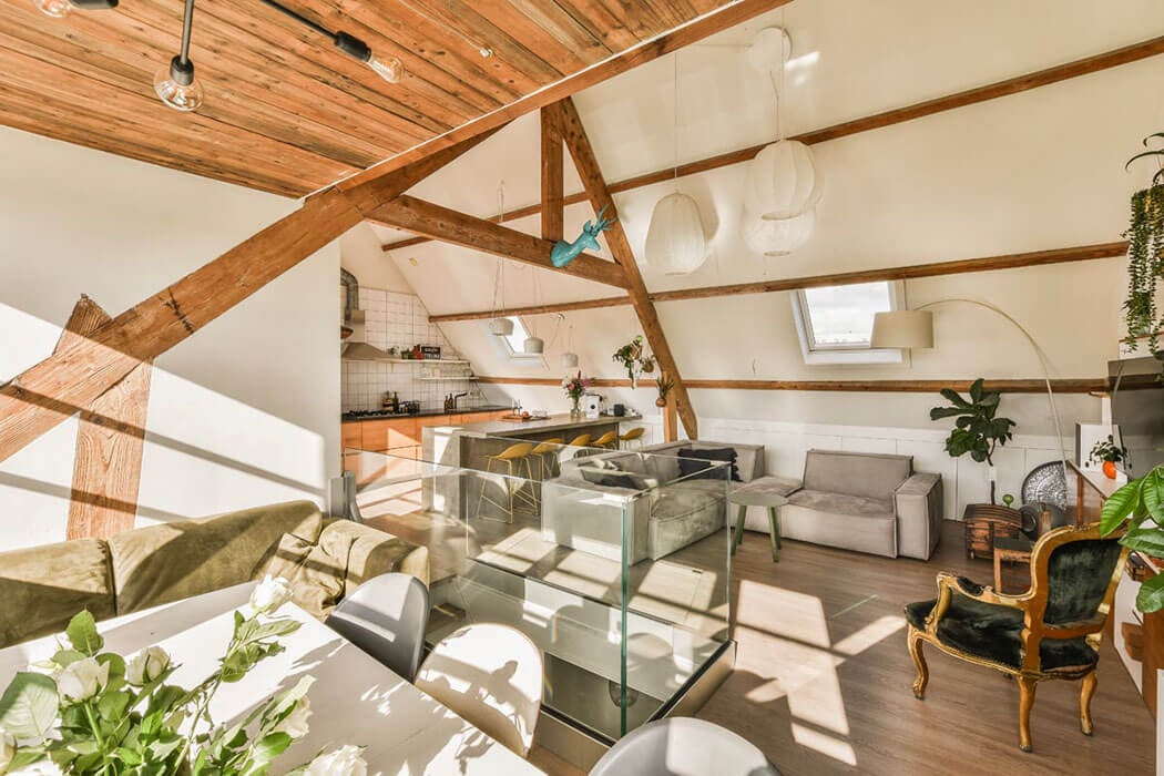 An open-plan attic apartment with exposed timber beams; on the left a dining table with white chairs and a flower arrangement, in the centre a glass balustrade, in the background an open kitchen with orange-fronted units, on the right sofas and a gilt armchair with a green velvet cushion.