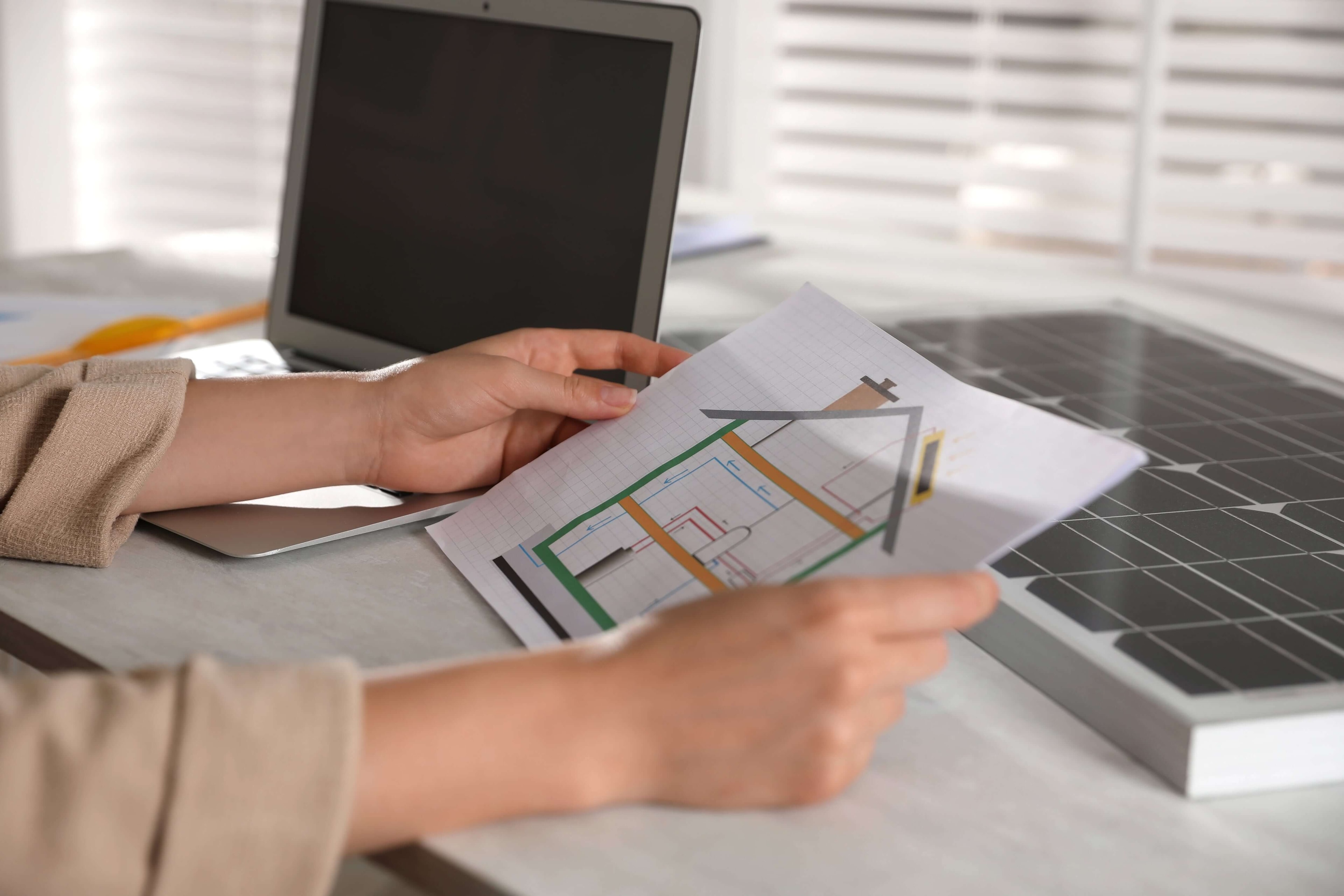 Two hands holding up a paper showing the cross-section of a three-storey house with a pitched roof and chimney, next to an open laptop and a drawing board for technical drawings.