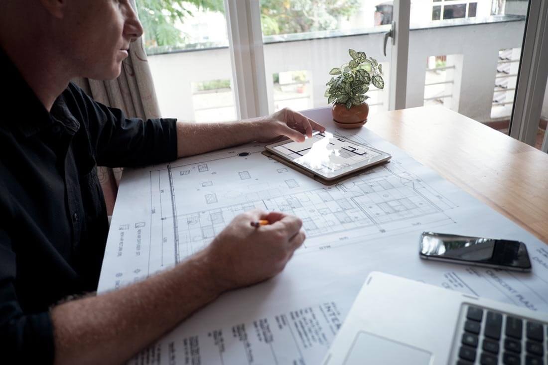 A man sitting at a wooden table, comparing a building plan in front of him with a floor plan visible on a tablet beside him.