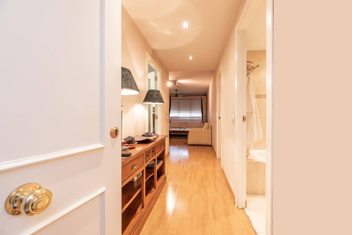View through the front door into a long, brightly lit interior hallway with several branching room entrances and on the right a sideboard with two identical table lamps on it.