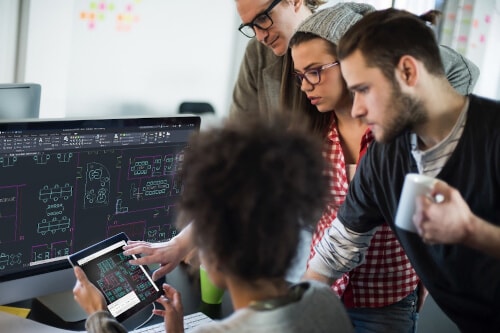 Several people standing around a desk with a PC workstation, looking at a floor plan on a tablet held up by the person with their back to the viewer.