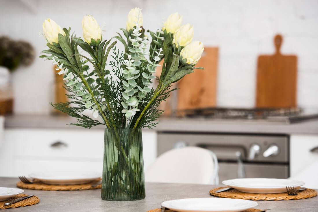Green glass vase with white flowers and eucalyptus branches on a set dining table with woven placemats and white plates; white kitchen blurred in the background.