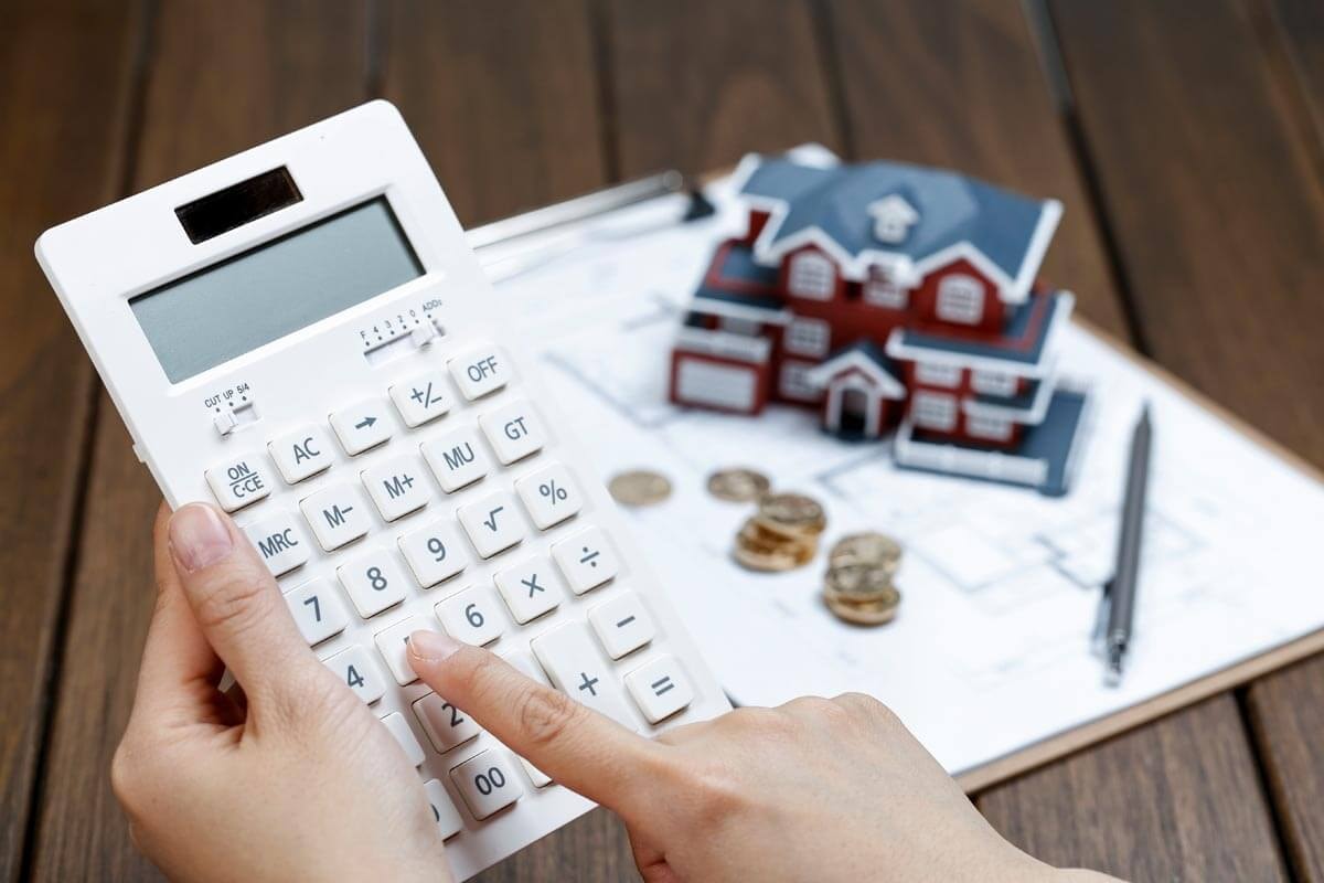 Close-up of hands holding a calculator, with a clipboard showing a floor plan and coins lying on it, and a spacious miniature house in the background