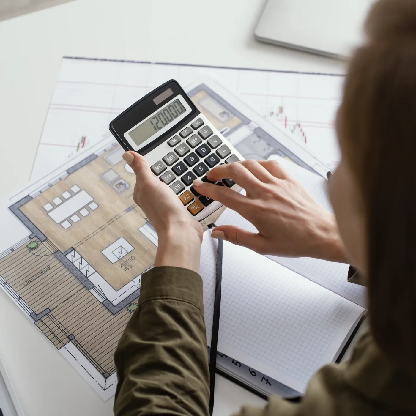 Rear view of a woman holding a calculator over colorful floor plans on a table, entering 120000