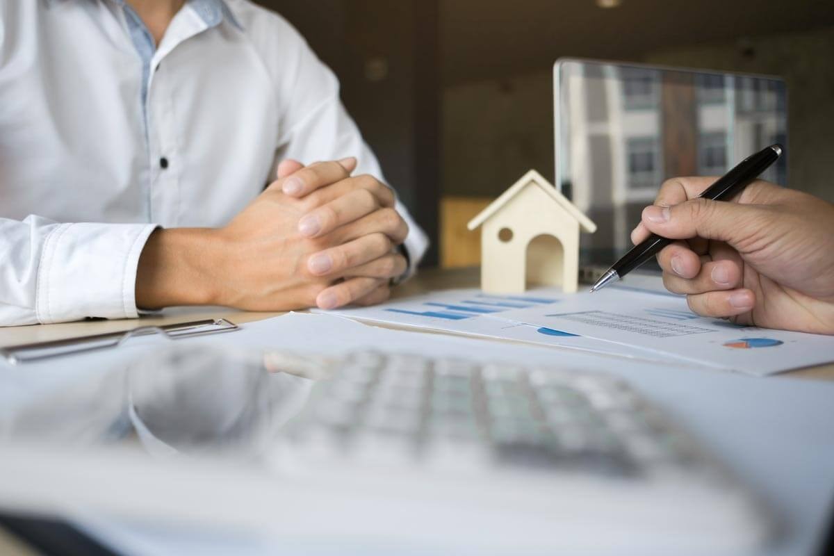 A financial expert sitting across the desk from another person, explaining a chart lying between them and pointing to its details with a ballpoint pen.