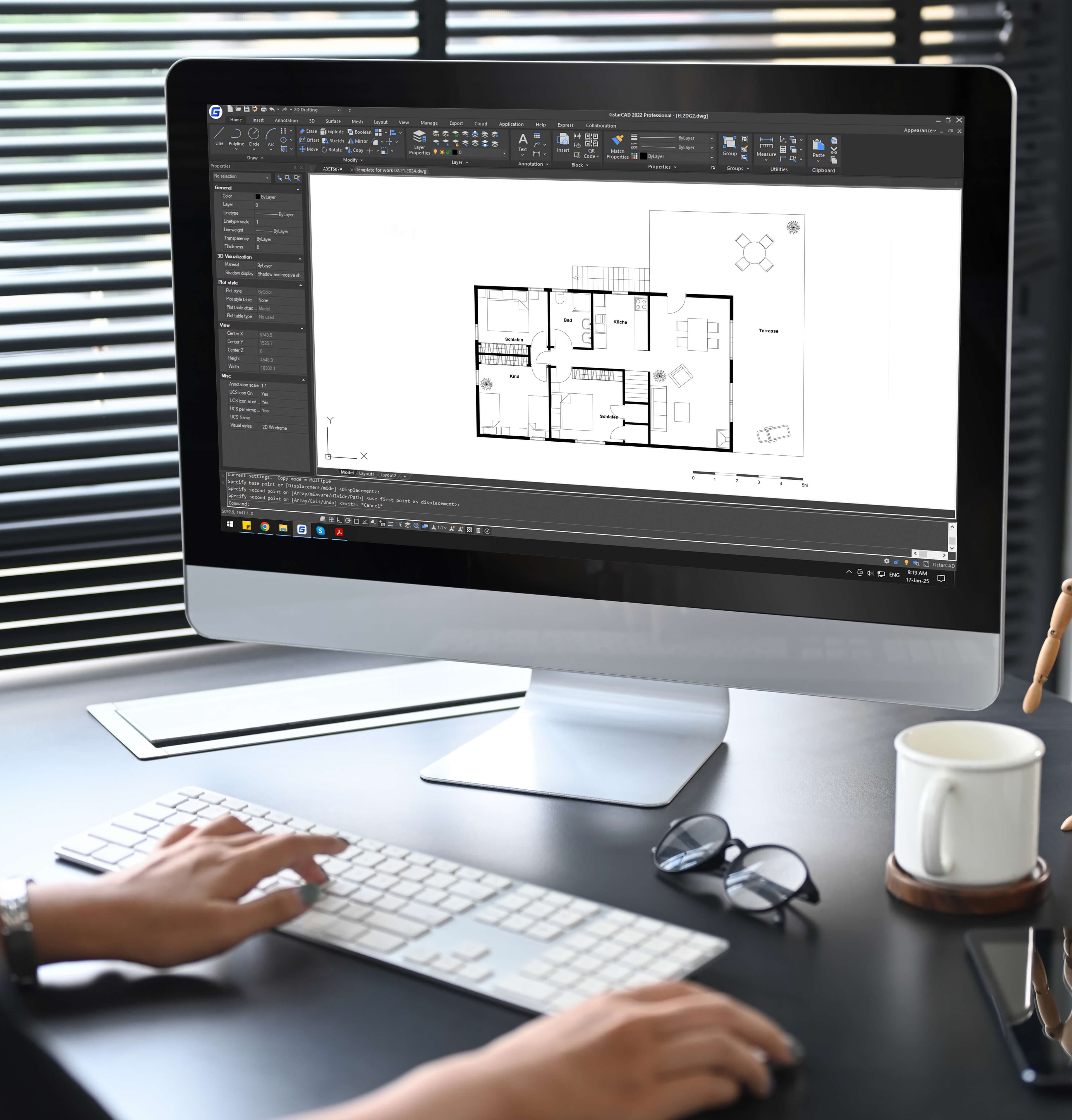 A person works with CAD software on a detailed floor plan with labelled rooms, with a keyboard, glasses and coffee mug on the desk beside them.