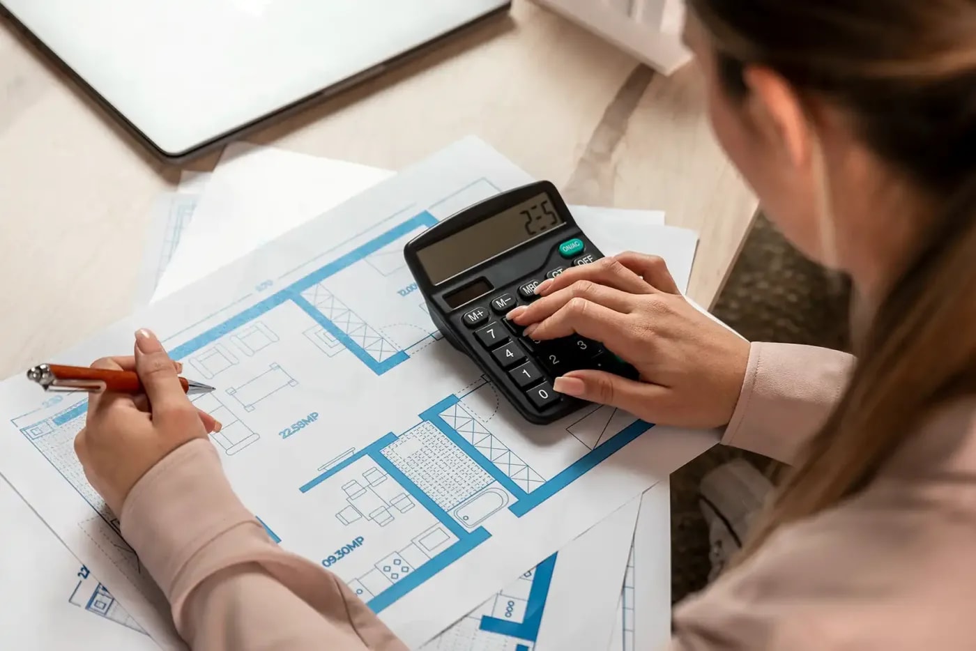 View over the shoulder of a woman sitting in front of printed floor plans, adding up the room areas on a calculator