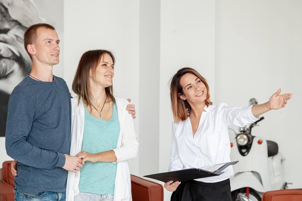 A smiling young couple stands arm in arm next to a female estate agent in her office, listening as she explains something she is pointing to, which is out of frame.