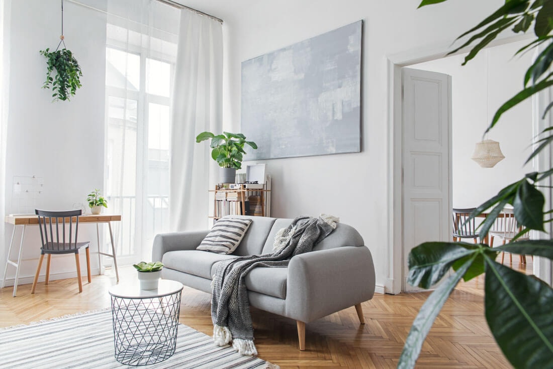 Bright, Scandinavian-style living room with a grey sofa, knitted throw blanket, wire coffee table, large abstract canvas and indoor plants on herringbone parquet.