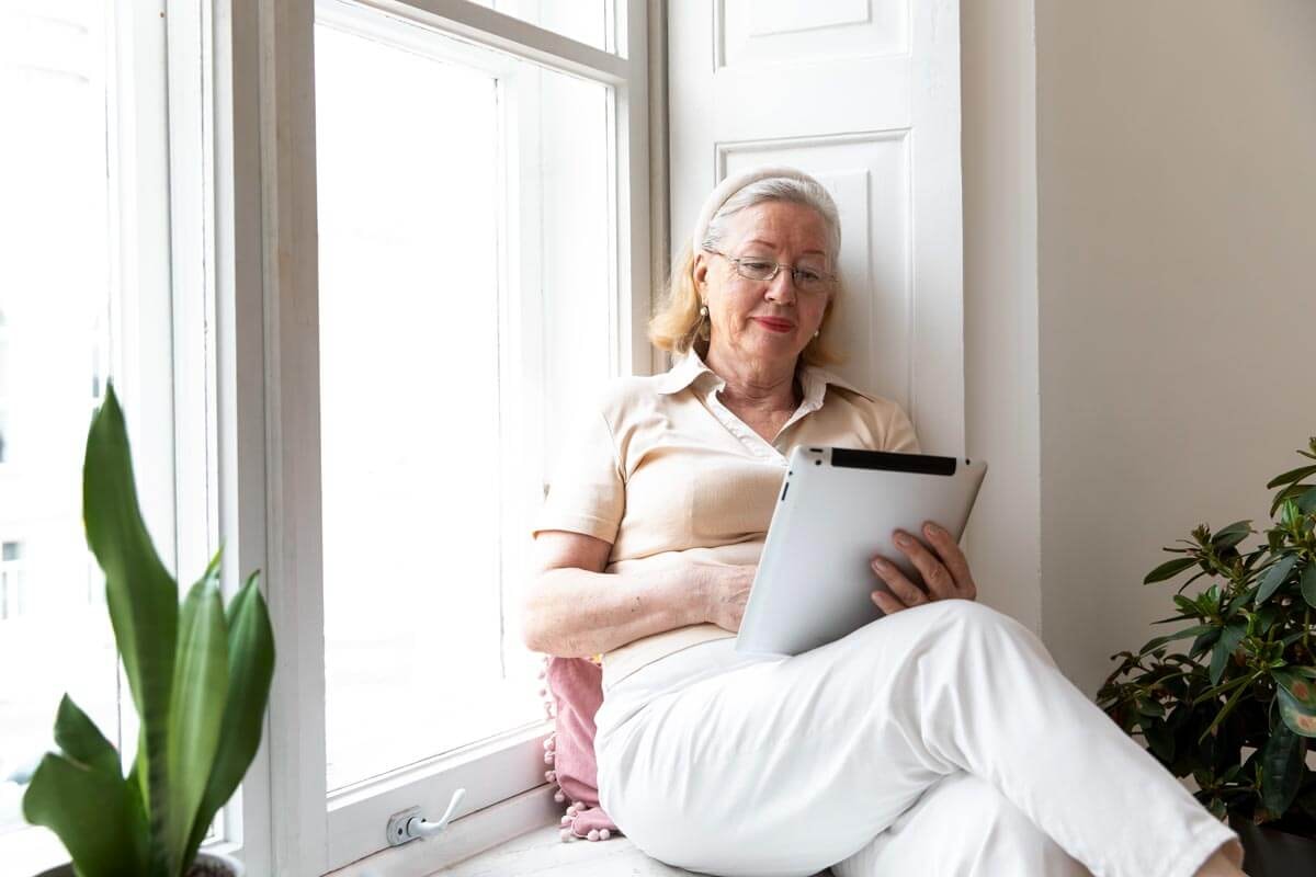 An elderly woman sits on a windowsill with her legs crossed, holding a tablet and reading from it.