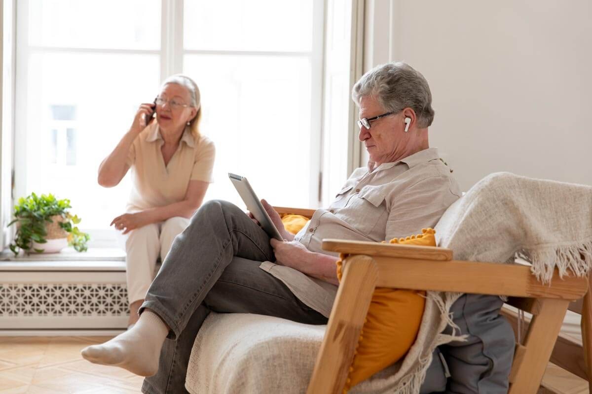 Two seniors in their living room — a man reading on the sofa in the foreground, a woman on the phone by the window in the background.
