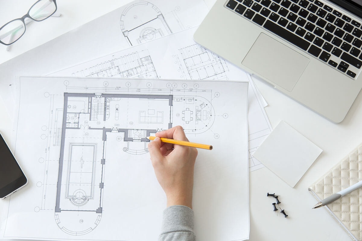 Top-down view of a desk covered in floor plans, a laptop and a pair of glasses, with a hand holding a pencil making notes on the topmost floor plan.