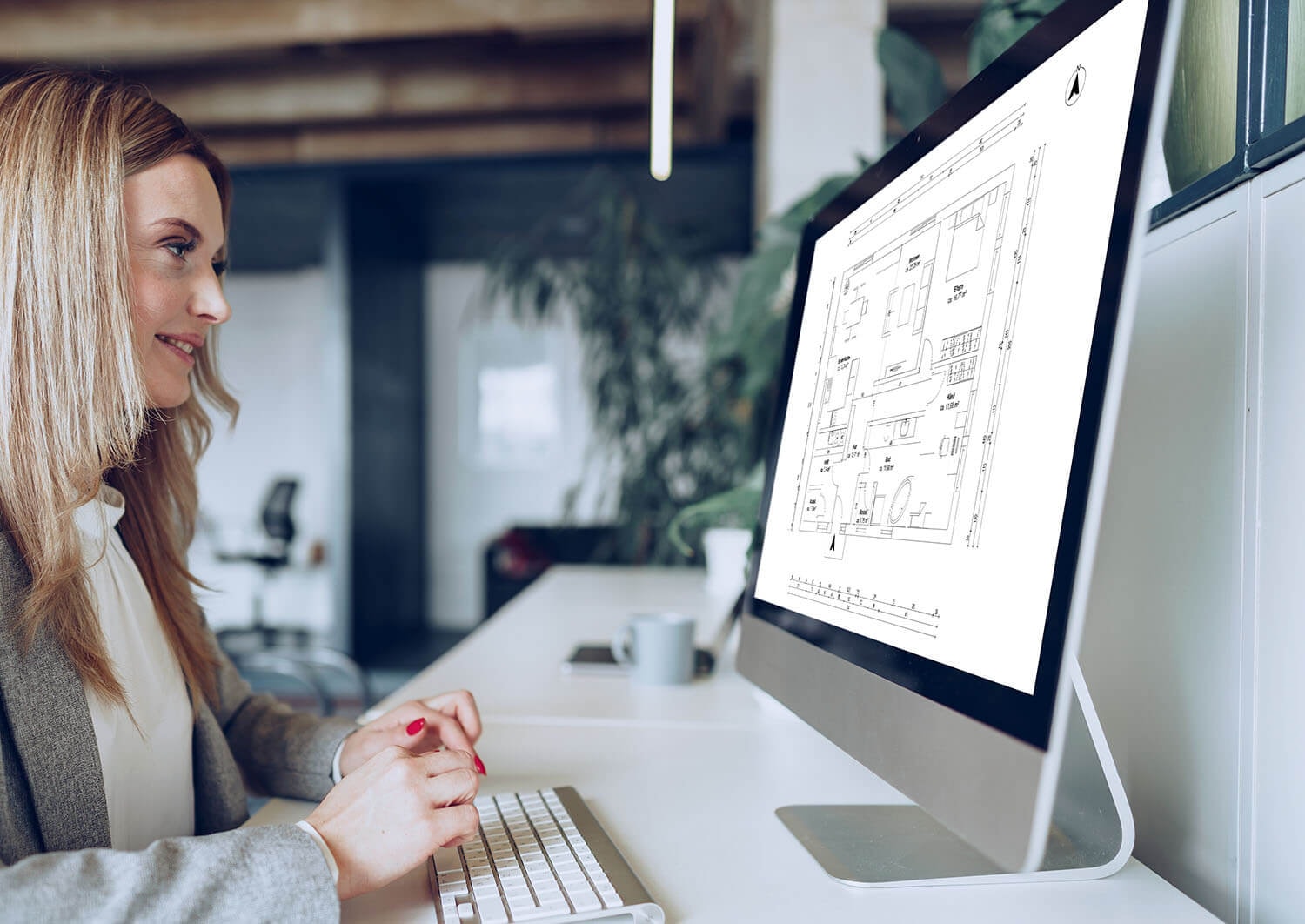 A woman sits at a desk in front of a PC editing the floor plan of a 3-room apartment with a combined living and dining area and an open kitchen