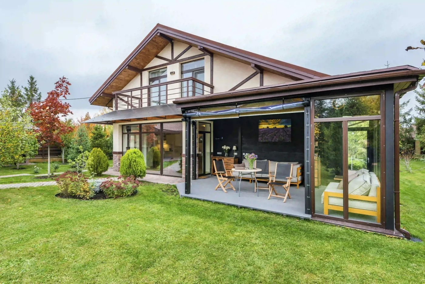 Blick von einer Gartengrünfläche auf ein modernes Einfamilienhaus im Retro-Fachwerklook mit flachem Satteldach, Wintergarten und Balkon und Mischgehölz im Hintergrund.