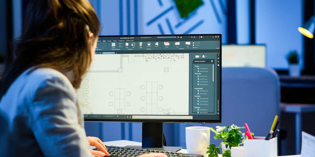 Rear angled view of a woman sitting in front of a large monitor displaying a floor plan in AutoCAD.