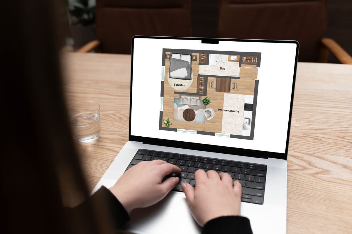 Over-the-shoulder view of a person at a wooden desk making changes to a floor plan on the laptop in front of them.